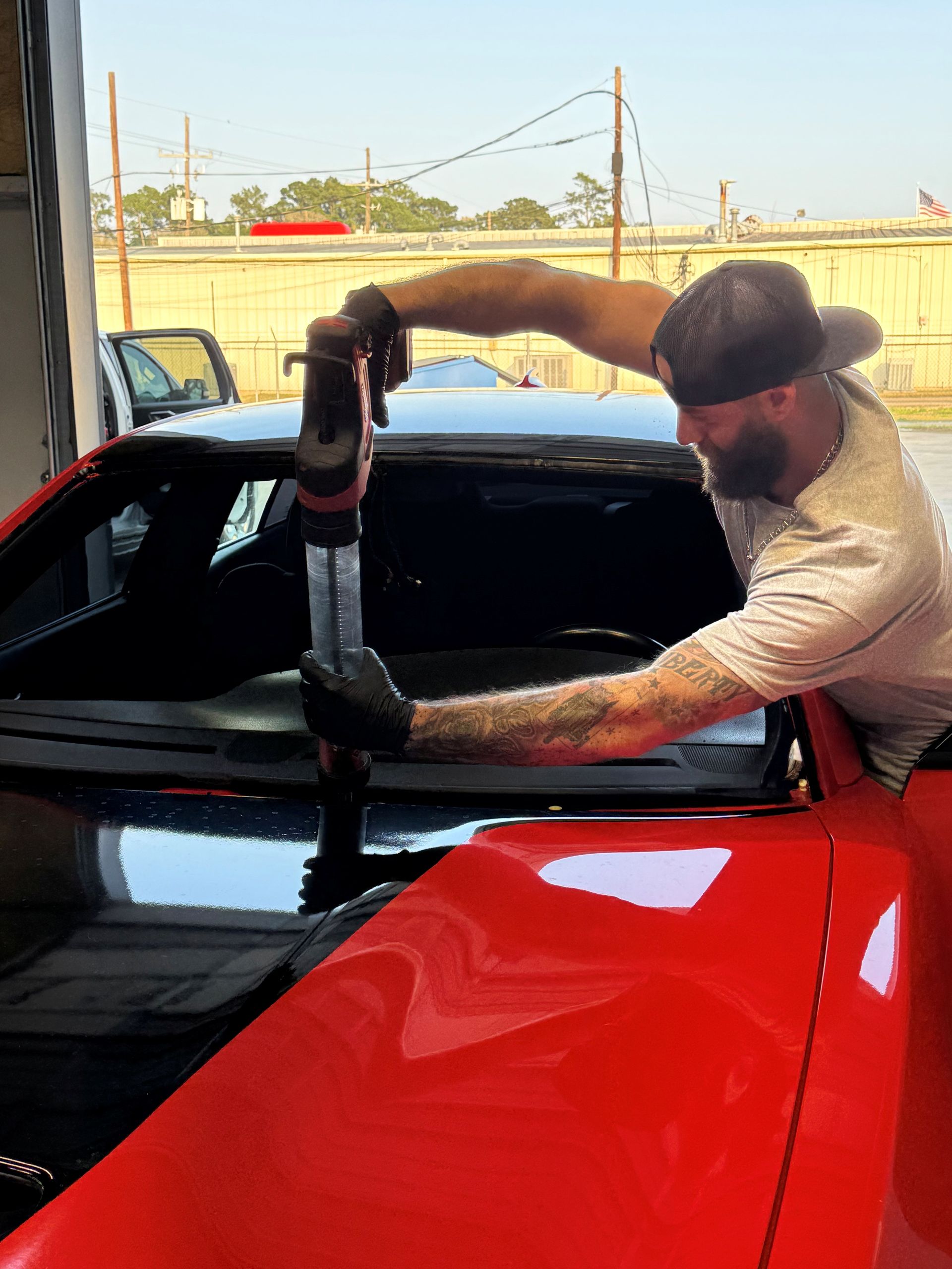 A person with a beard and tattoos uses a powered tool to apply adhesive to the windshield area of a bright red car.