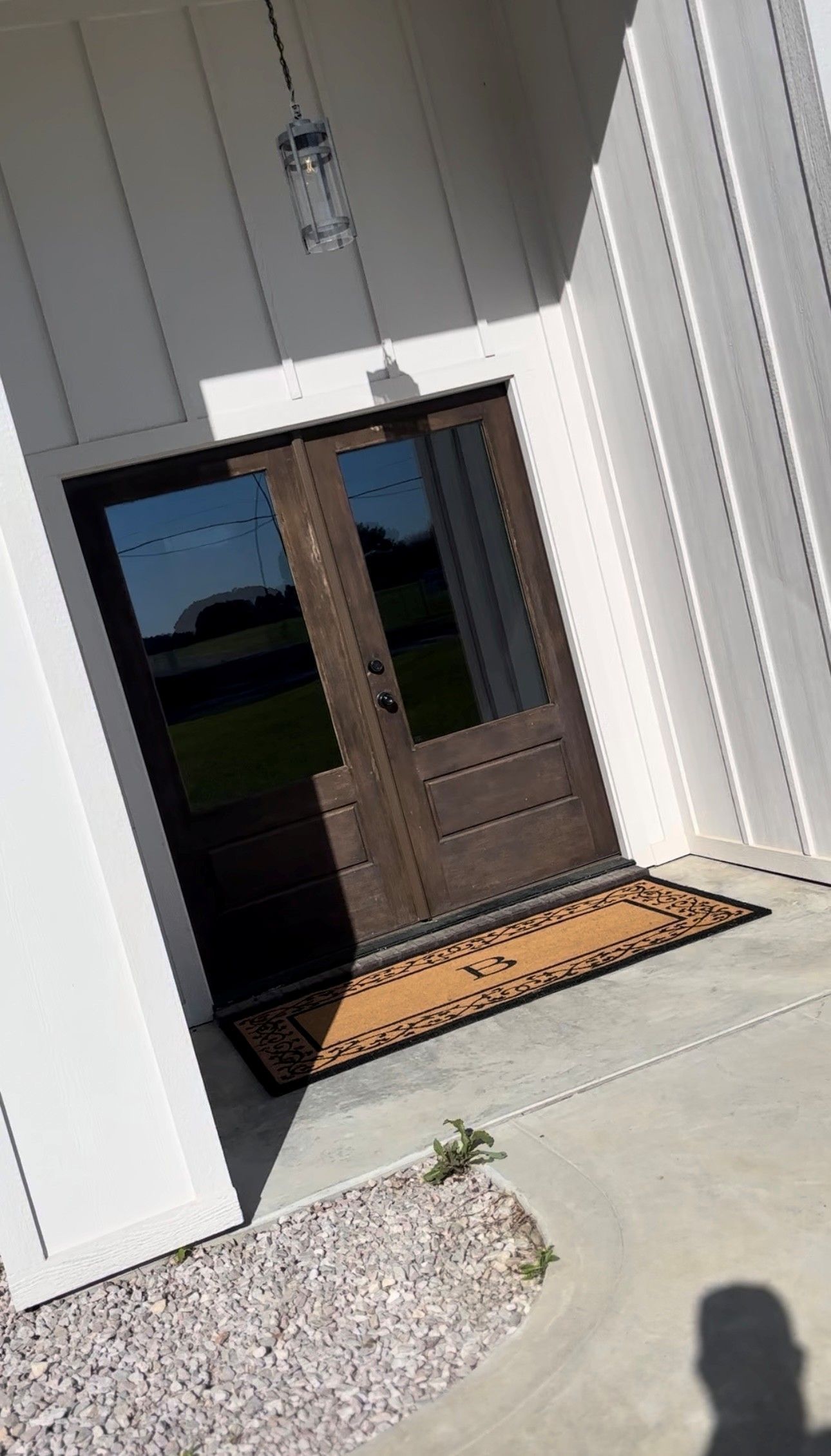 A front porch with double dark wood doors, a welcome mat, and a hanging pendant light against white board-and-batten siding.