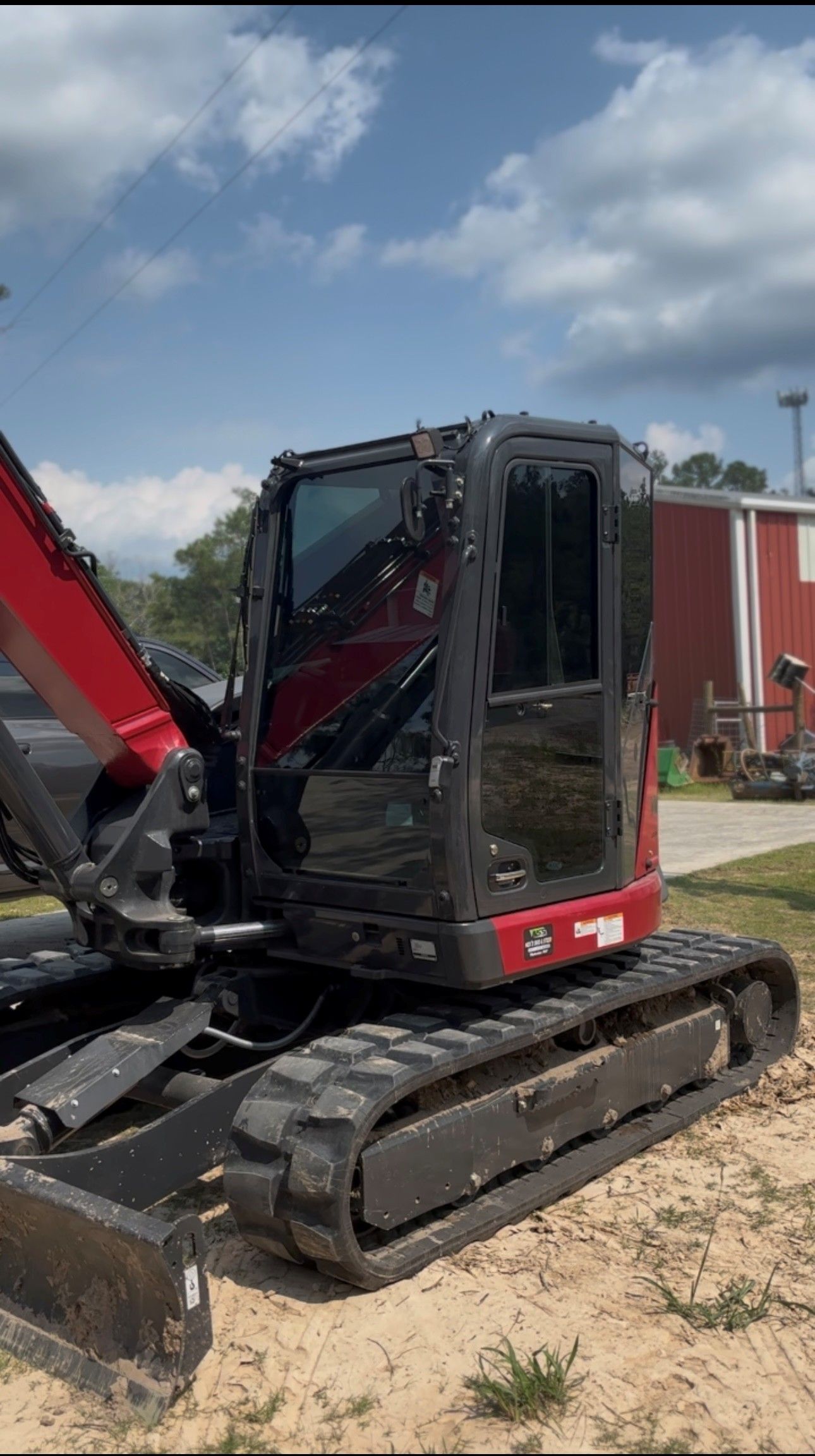 A red Takeuchi compact excavator with a closed cab parked on dirt against a blue sky with clouds.
