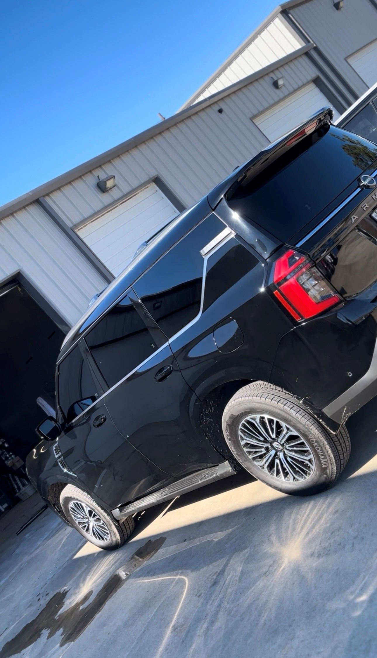 A black SUV parked at an angle in a concrete lot in front of a metal warehouse building under a bright blue sky.