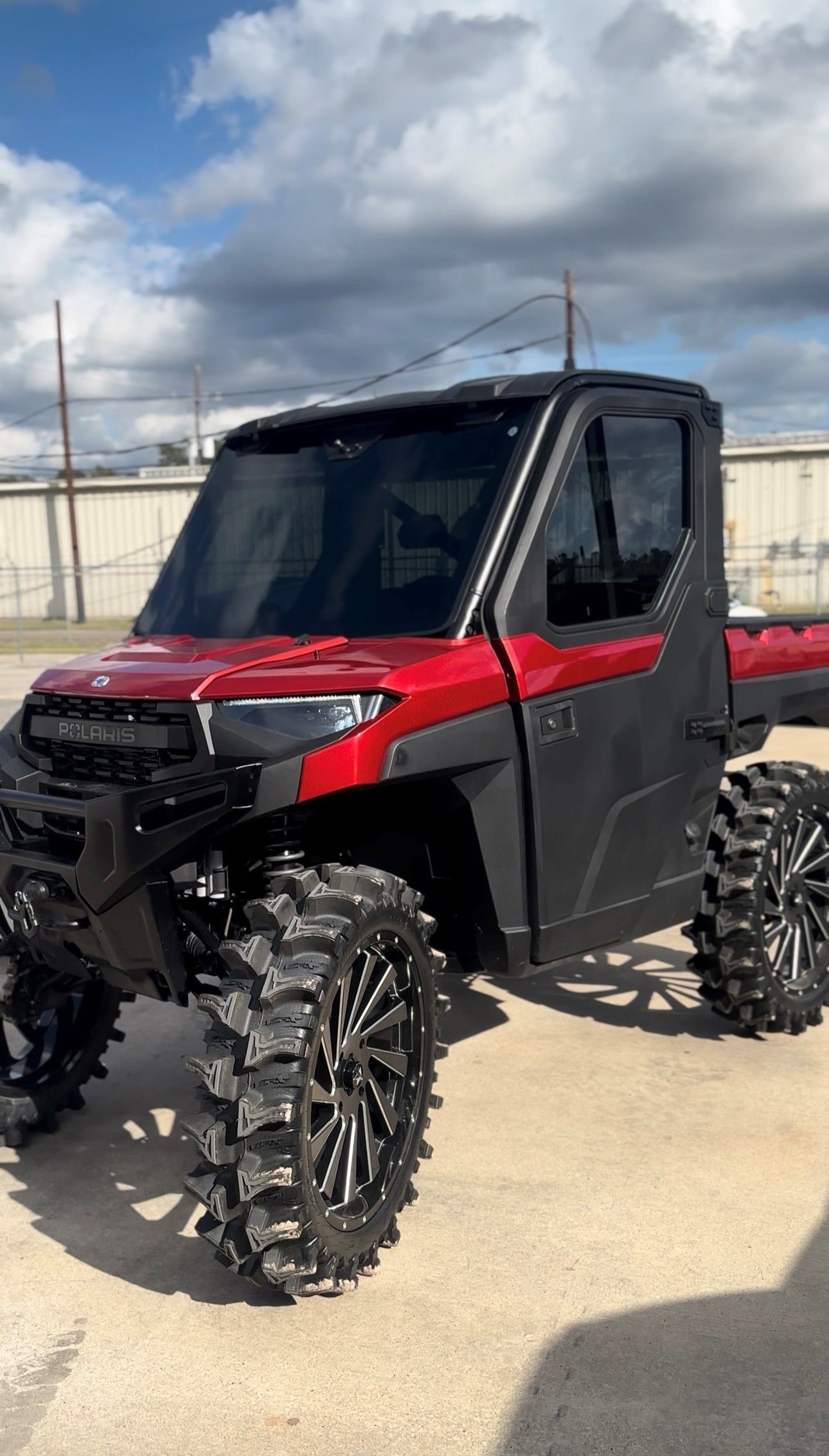 A red and black utility task vehicle with oversized mud tires parked outdoors under a cloudy sky.