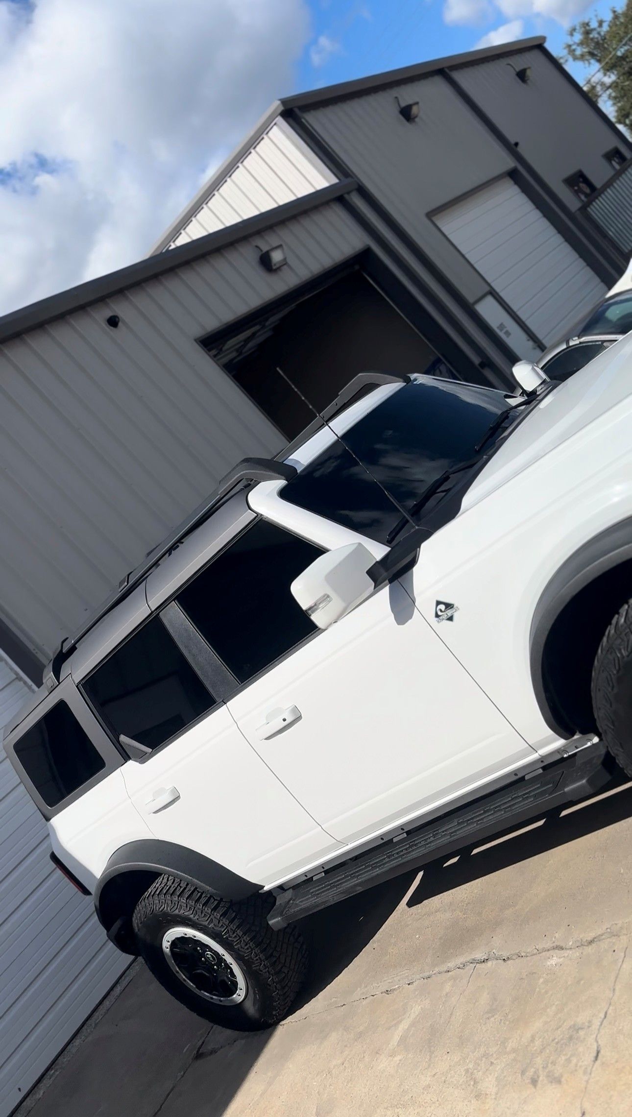 A white Ford Bronco parked on concrete in front of a gray metal building under a partly cloudy sky.