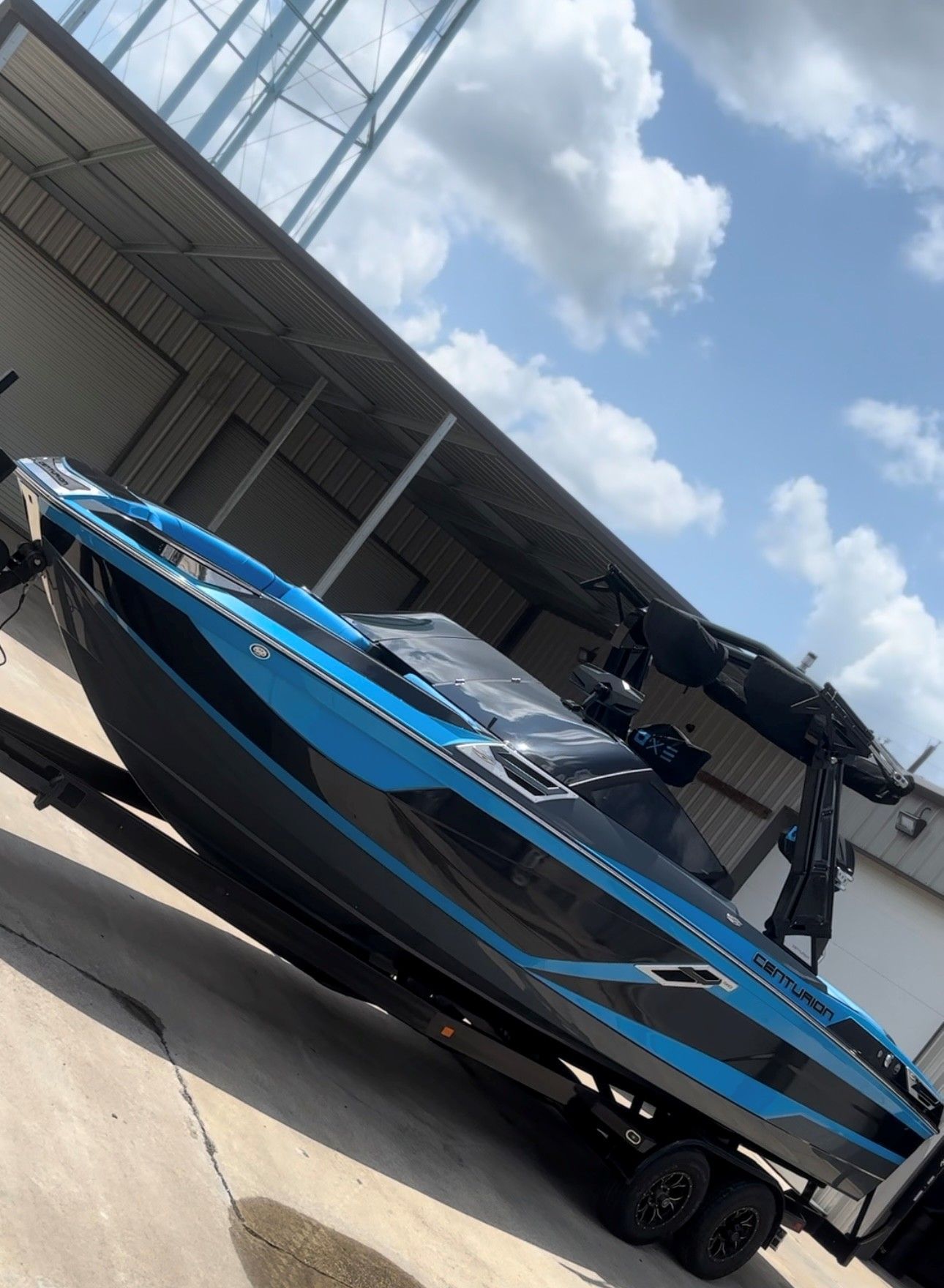 A blue and black motorboat sits on a trailer in a parking area under a metal awning on a sunny, cloudy day.