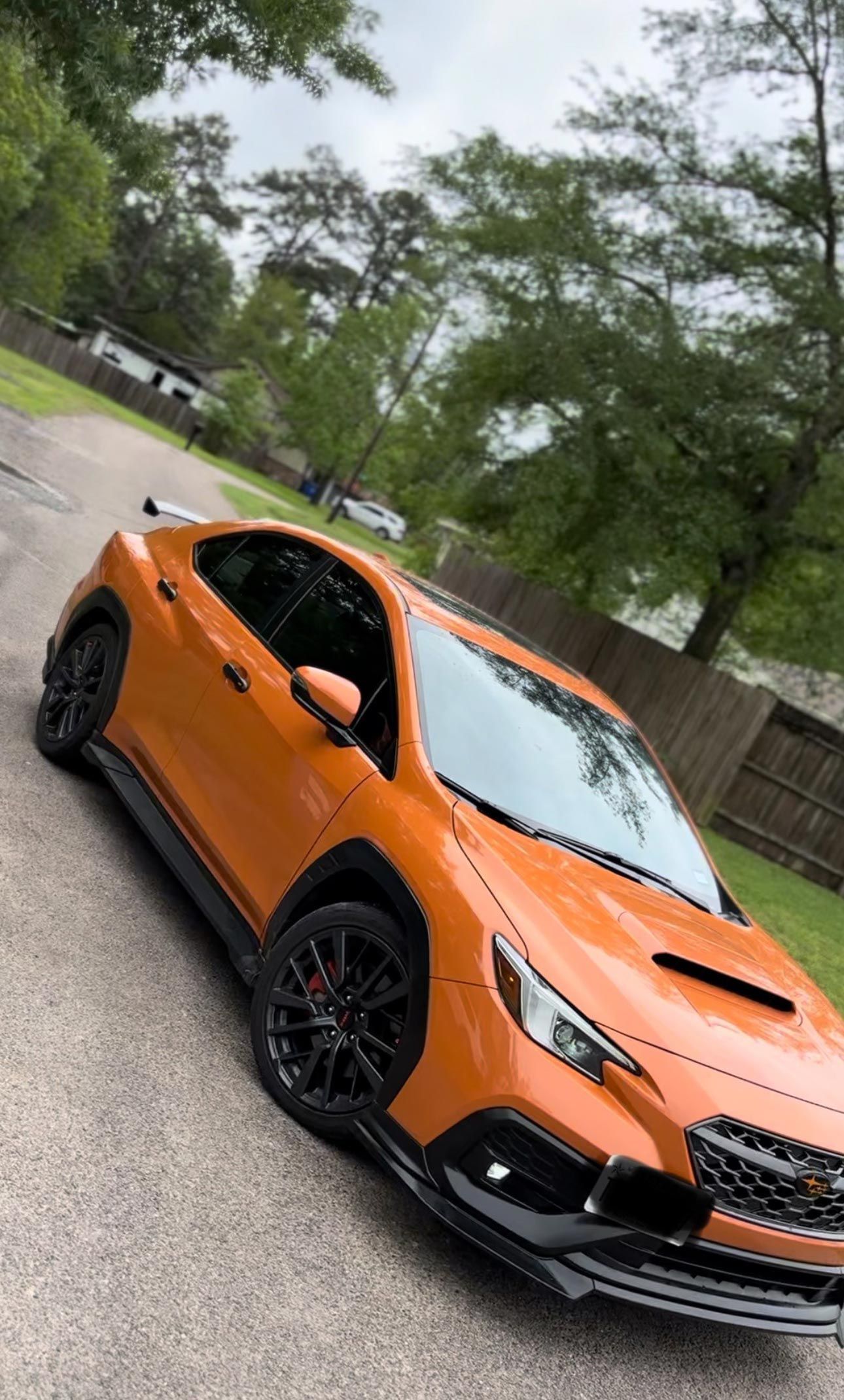 An orange Subaru WRX parked on a paved driveway surrounded by trees and a wooden fence.