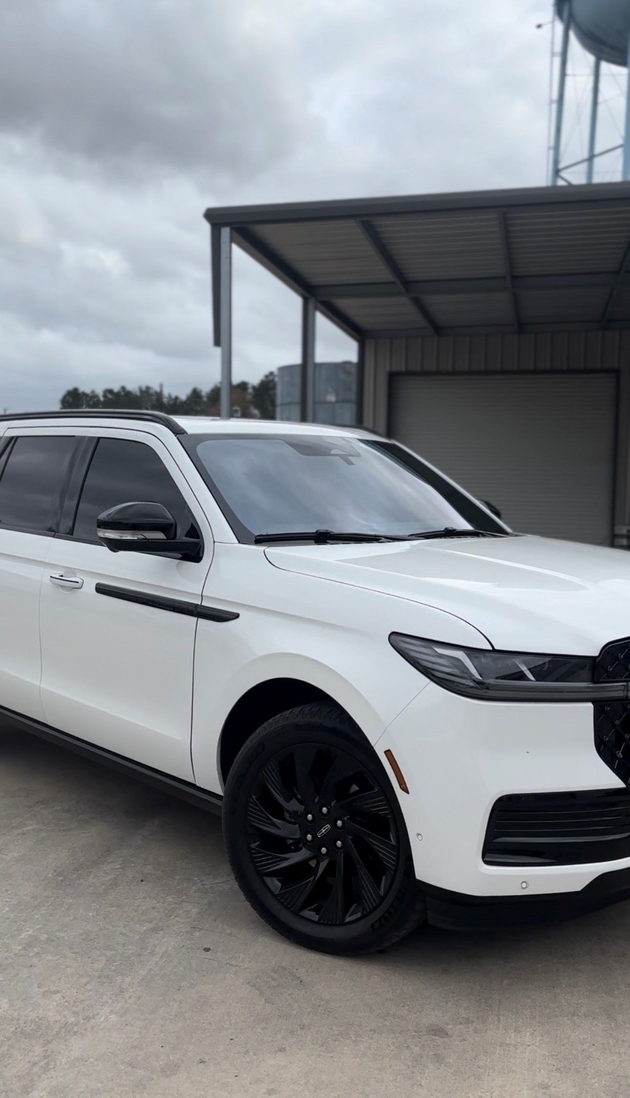 A white SUV with black wheels parked on a concrete lot next to a building and water tower under a cloudy sky.