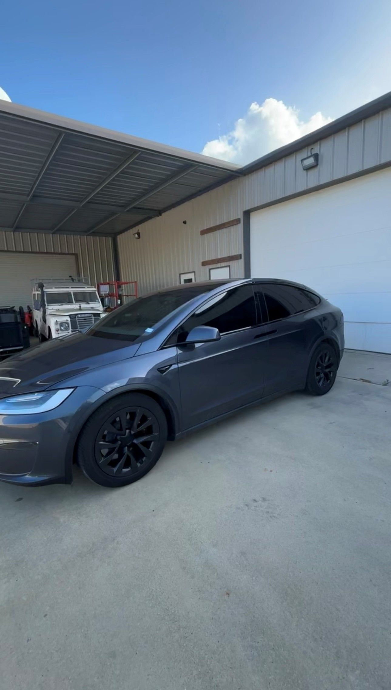 A dark gray Tesla Model X parked outside a workshop under a metal canopy against a clear blue sky.