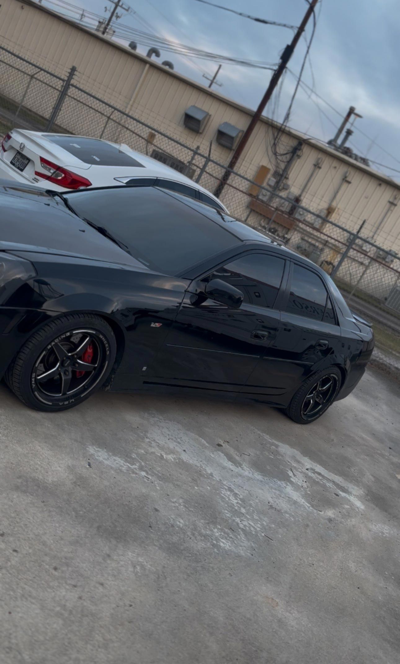A black sedan with tinted windows and black rims parked on a concrete lot, with a white car partially visible behind it.