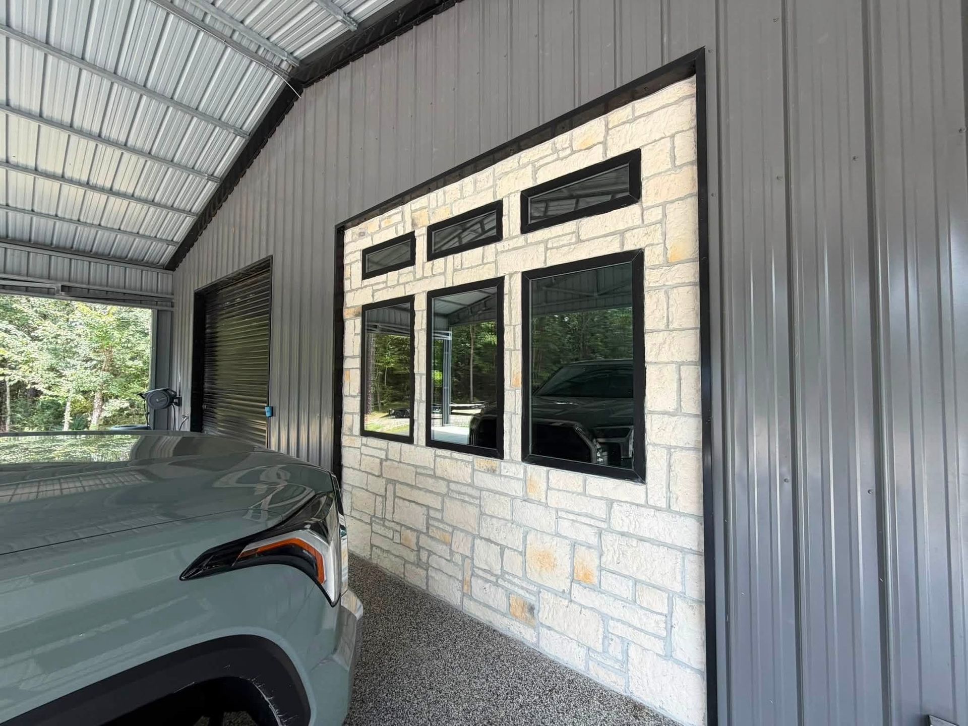Covered carport with a gray vehicle parked beside a windowed stone wall and corrugated metal siding