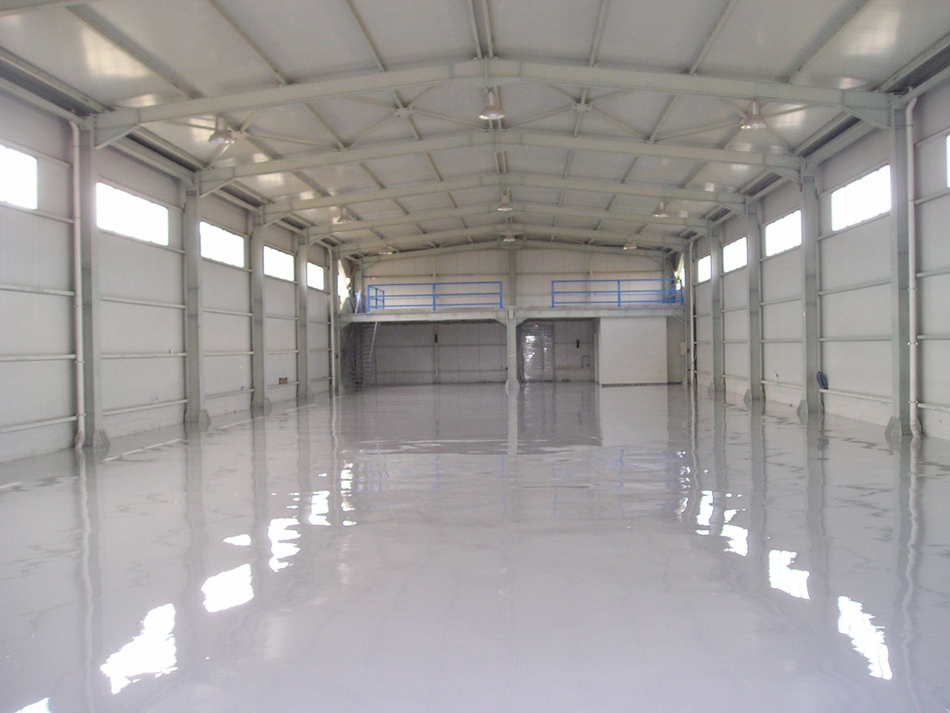 Empty, light-filled warehouse interior with reflective floor, metal beams, and windows.