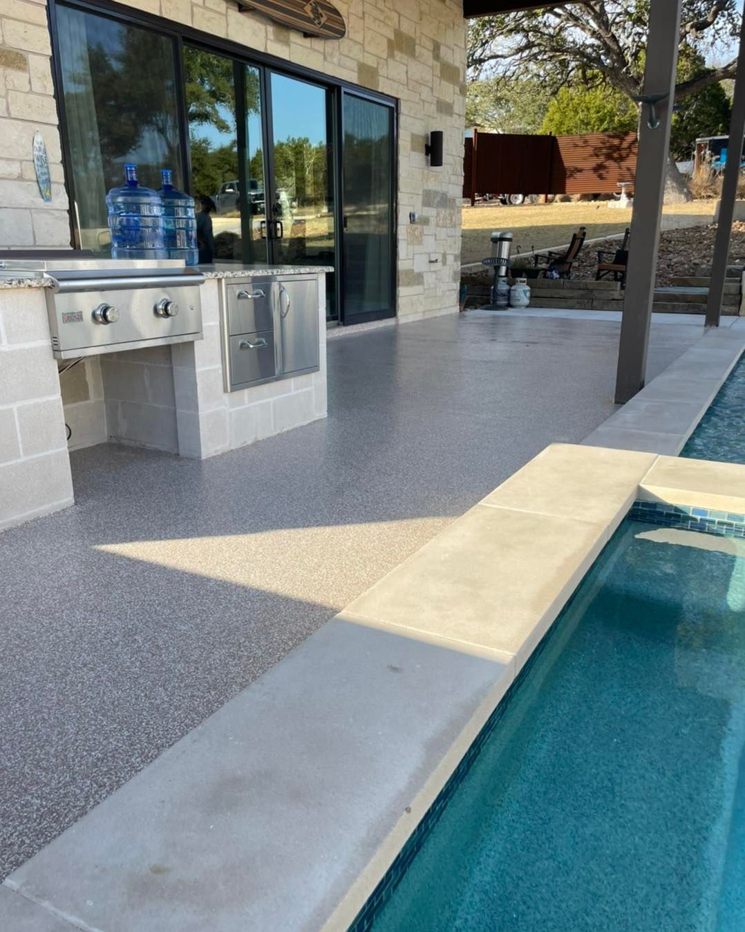 Outdoor kitchen with pool. Gray speckled concrete patio, stainless steel grill, and pool with blue water.