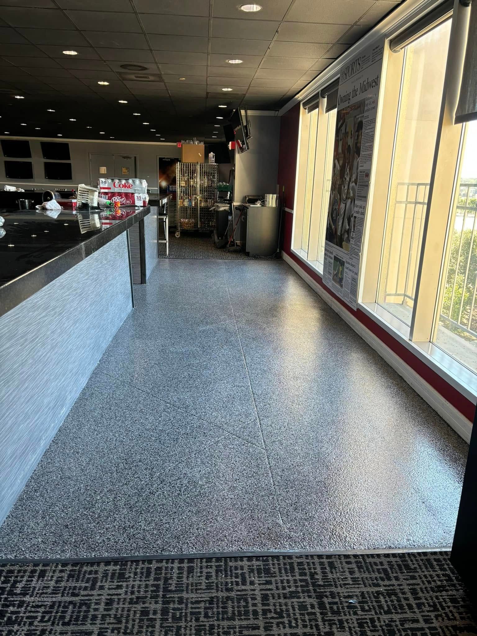 Interior shot of a bar. Gray flooring, black counter, and large windows with partially drawn blinds.