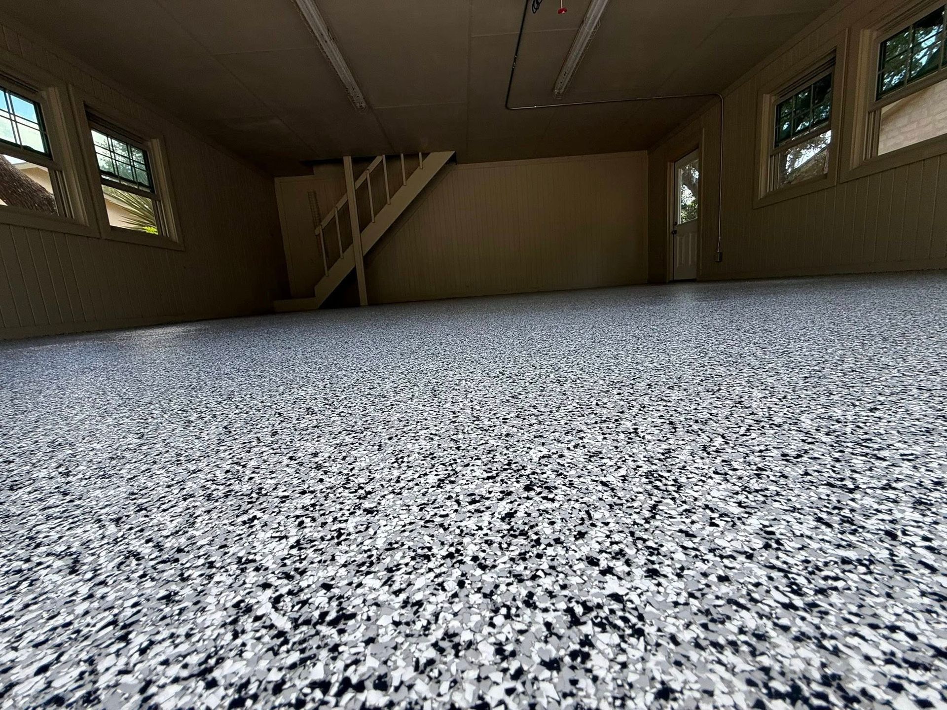 Garage interior with speckled gray floor, staircase, windows, and a door.