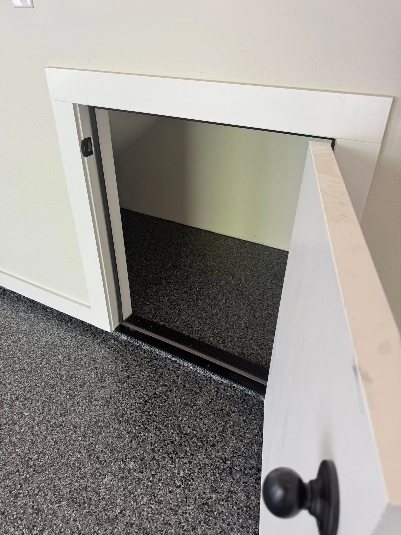 White door opens into a small storage space, black hardware. Dark speckled flooring and white walls.