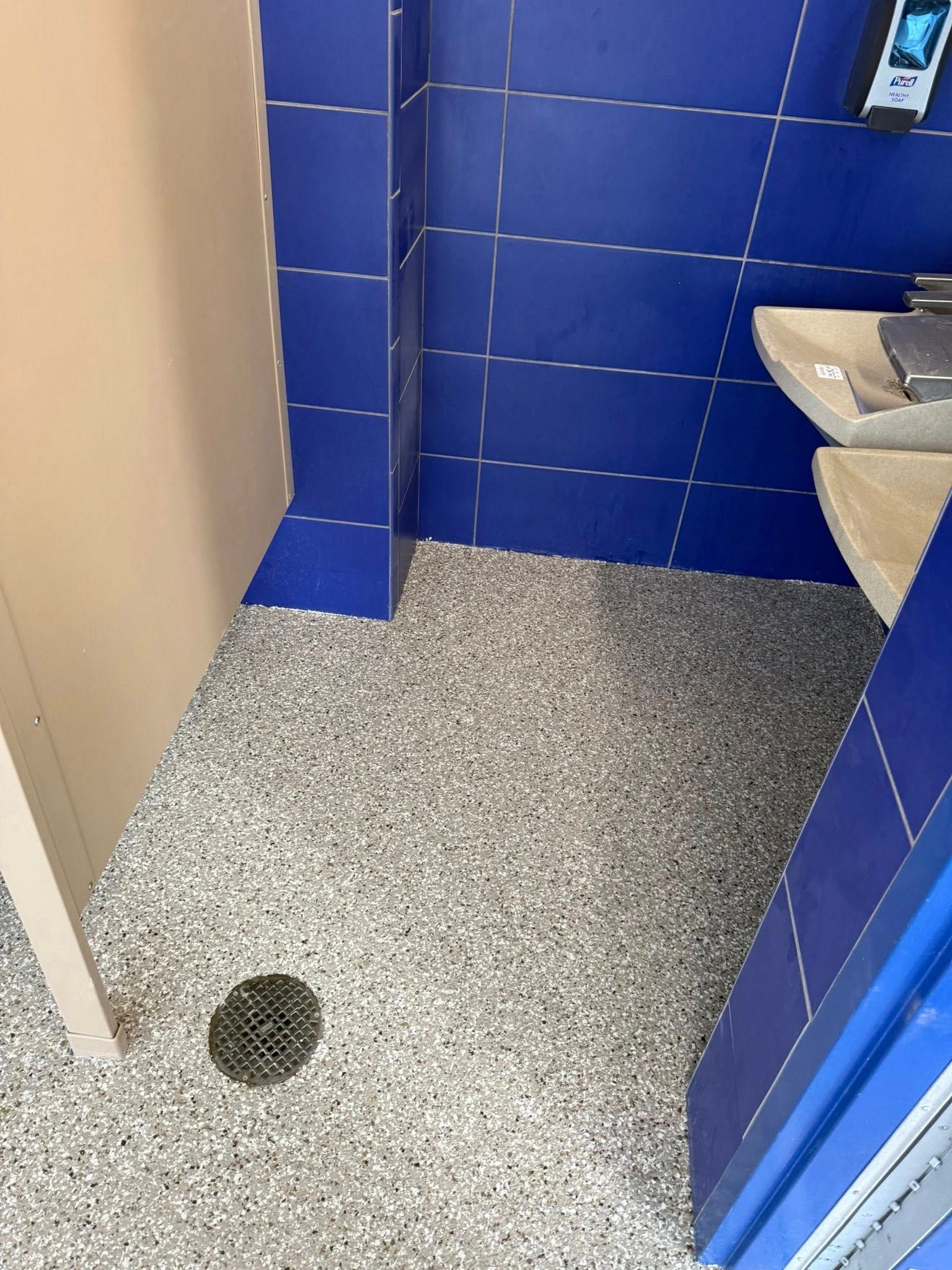 Public restroom with speckled flooring, blue tiled walls, and beige stall.