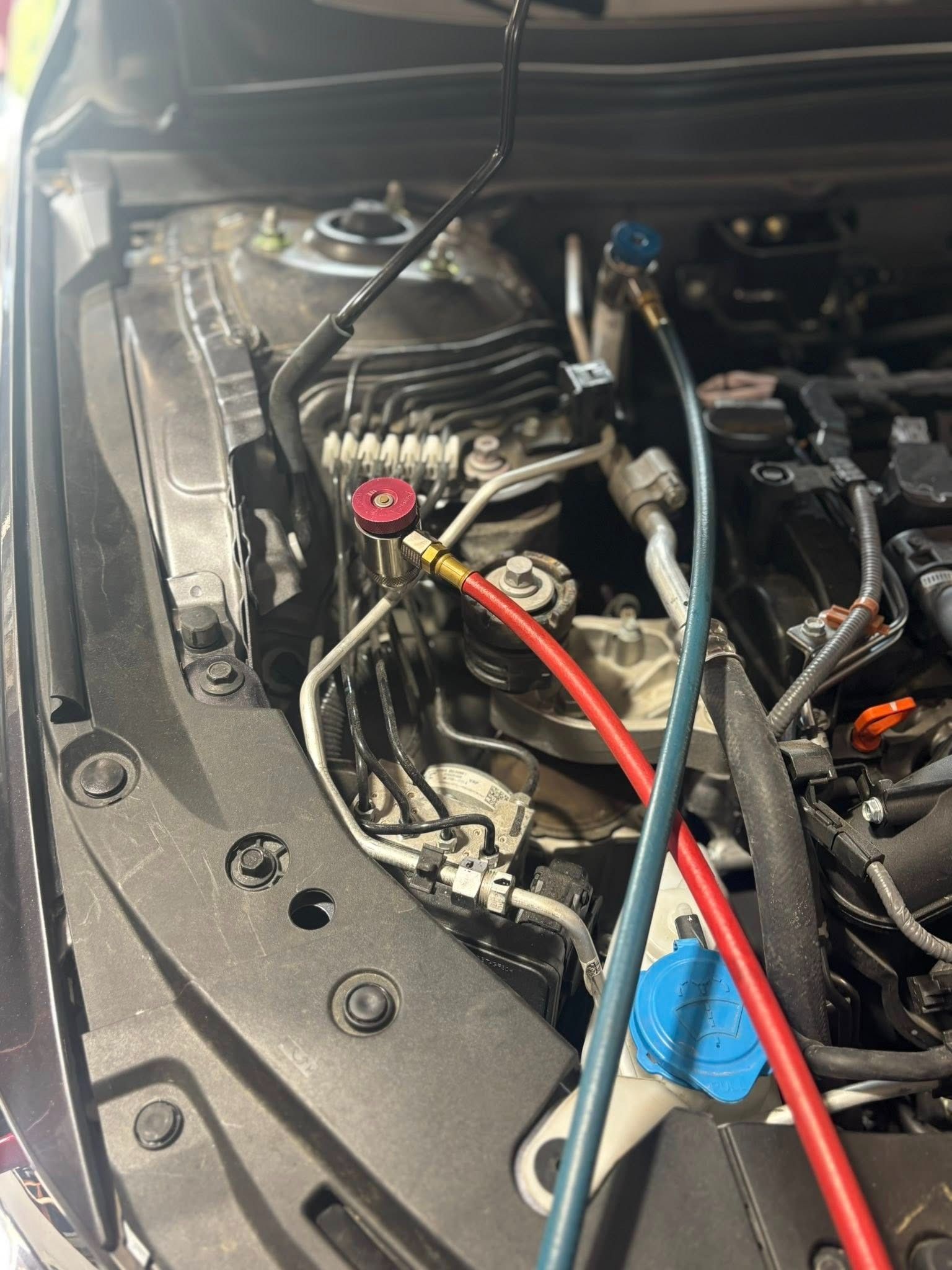 Car engine bay with AC lines connected. Red and blue hoses attached to valves, likely for refrigerant servicing.