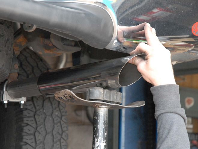 Person installs black exhaust tip on a vehicle's exhaust pipe, likely in a garage.