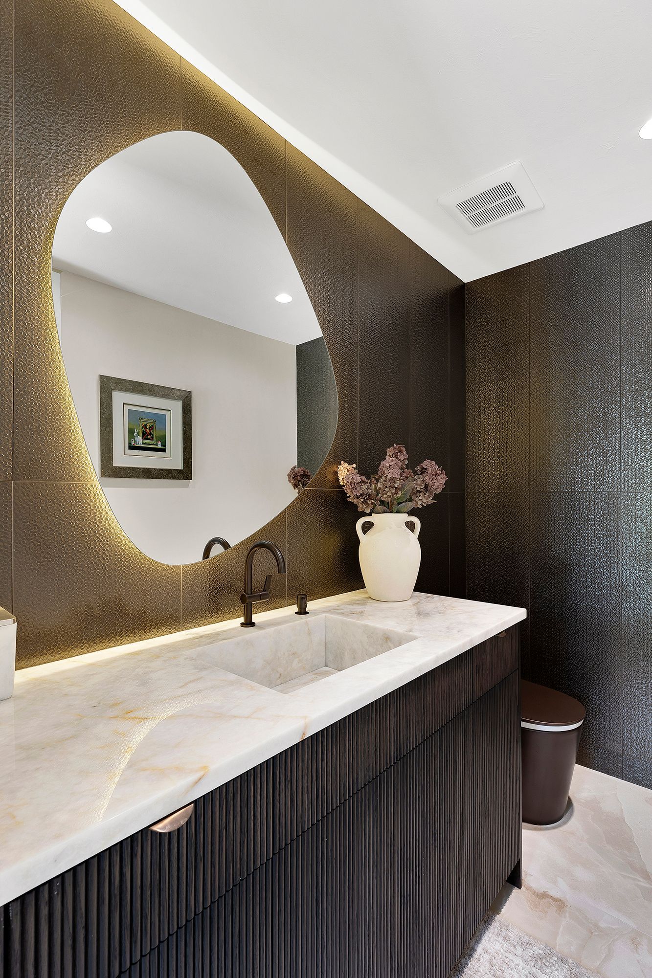 Modern bathroom vanity with oval mirror, marble countertop, dark textured walls, and white vase decor