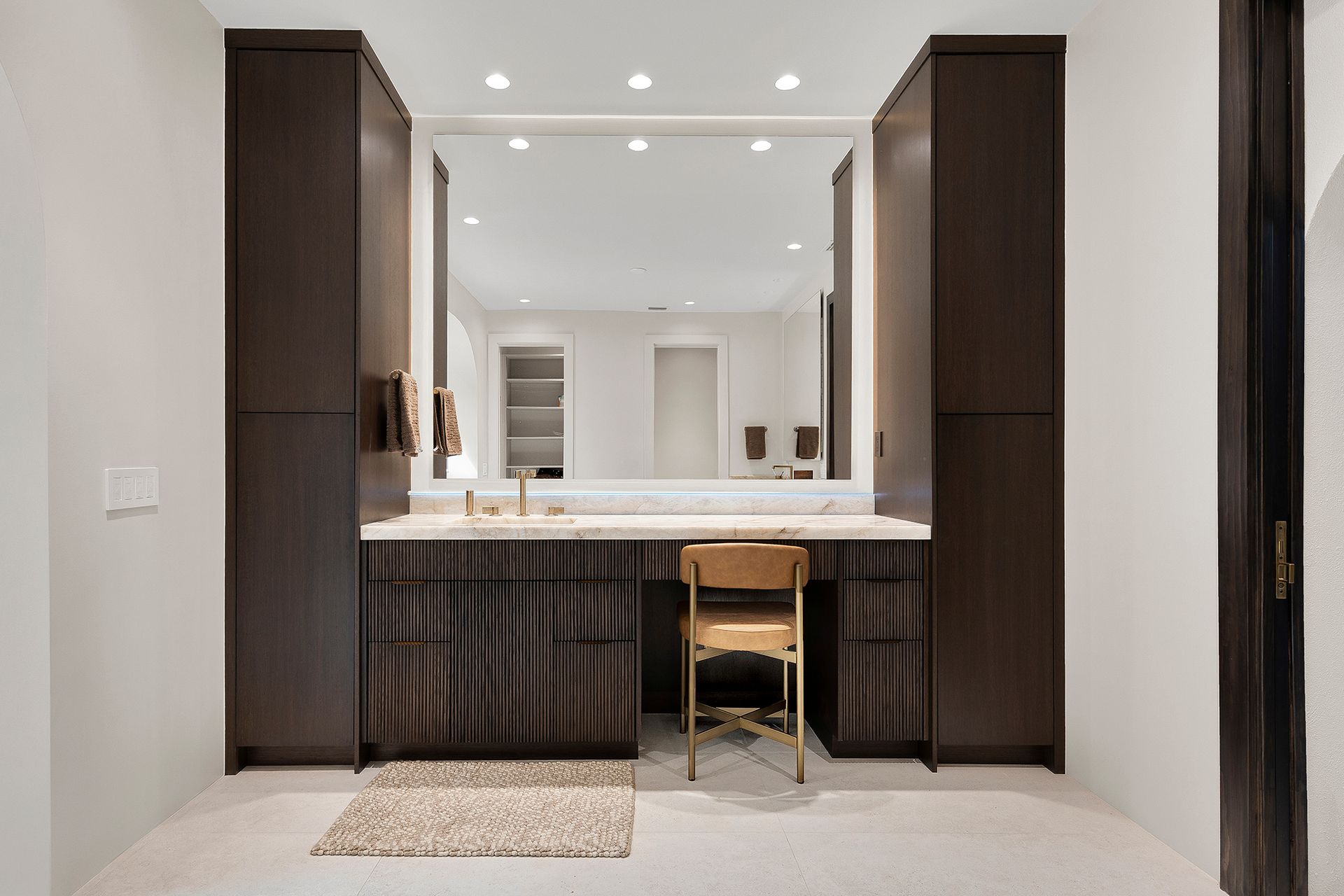 Modern vanity with large mirror, dark wood cabinets, white countertop, and a stool in a bright bathroom