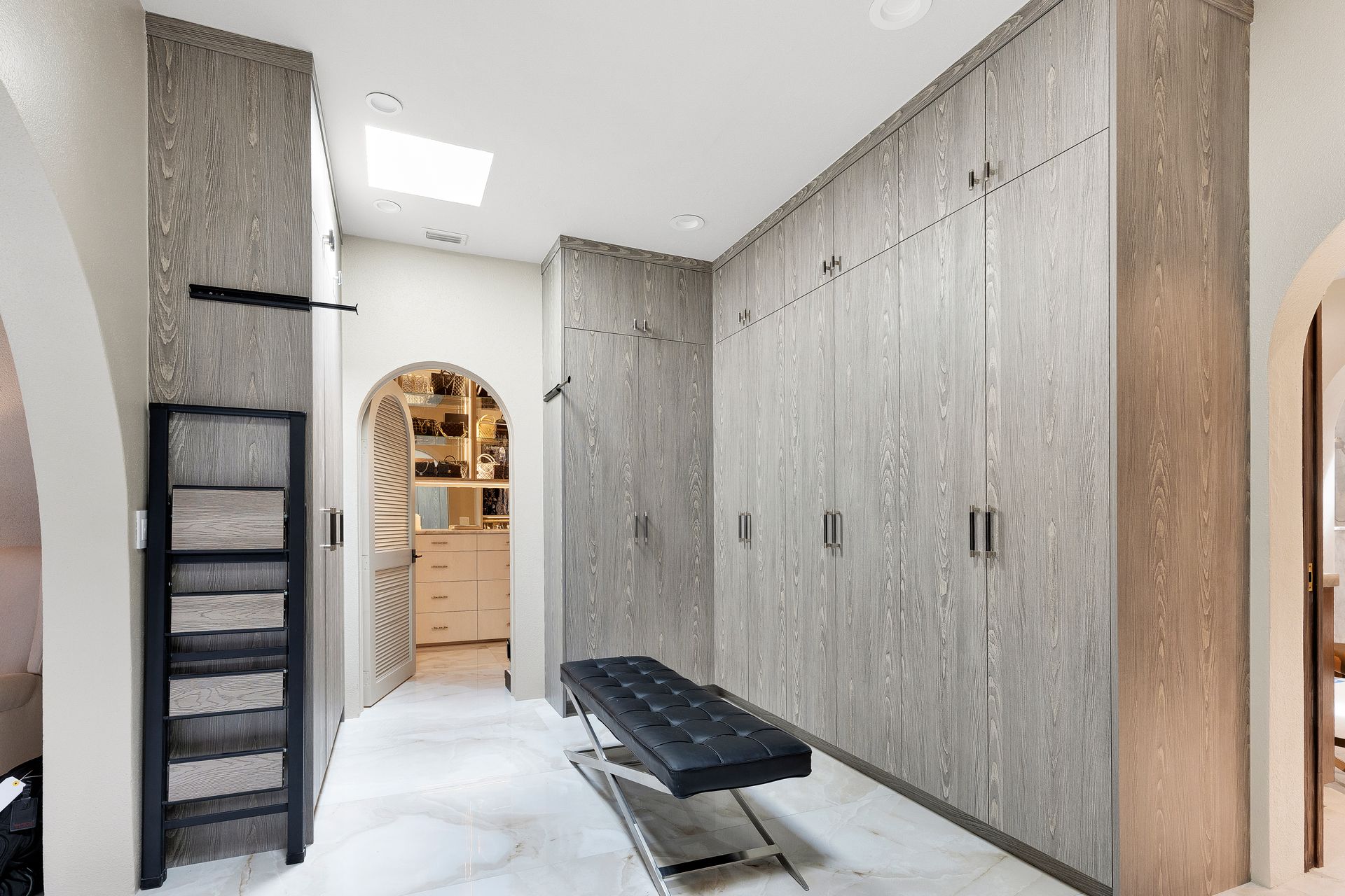 Bright walk-in closet with gray built-in cabinets, black bench, ladder shelf, and arched doorway to another room.