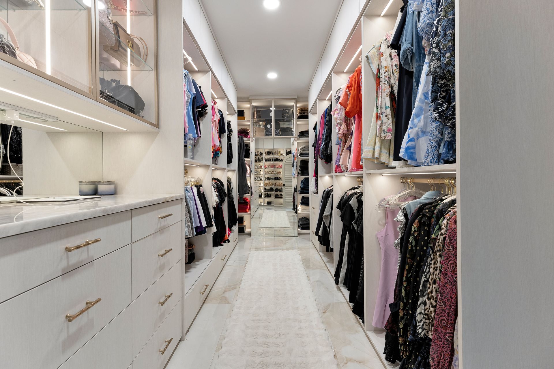 Bright walk-in closet with white shelves, drawers, and hanging clothes in a long, narrow aisle.