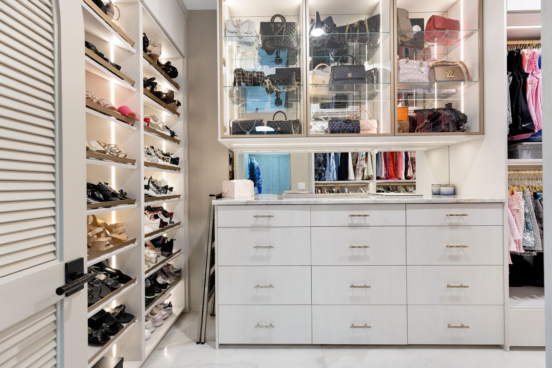 White walk-in closet with shelves of shoes, glass-front cabinets, and a large white dresser.
