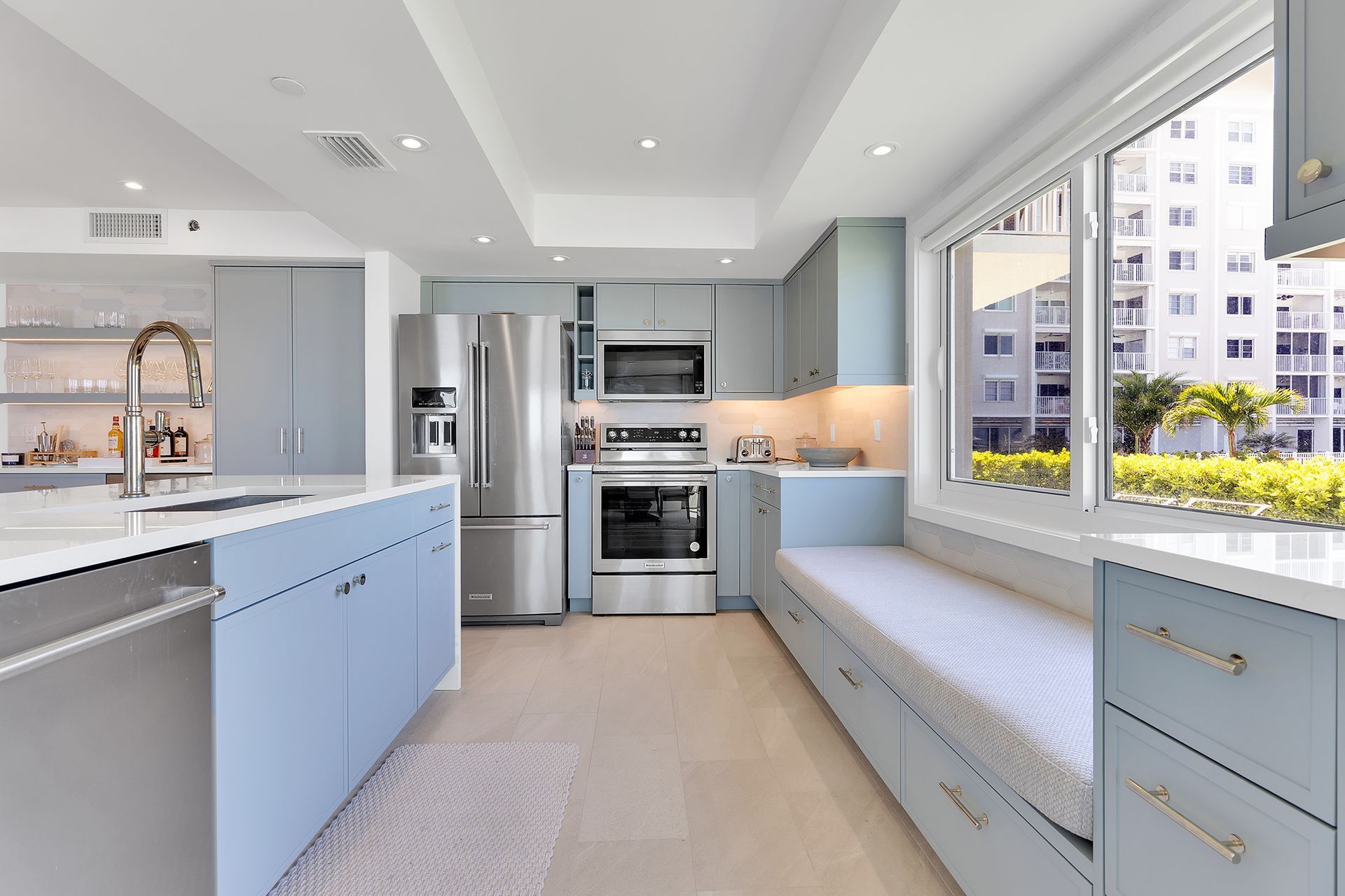 Bright modern kitchen with blue cabinetry, stainless appliances, and a long window seat by large windows