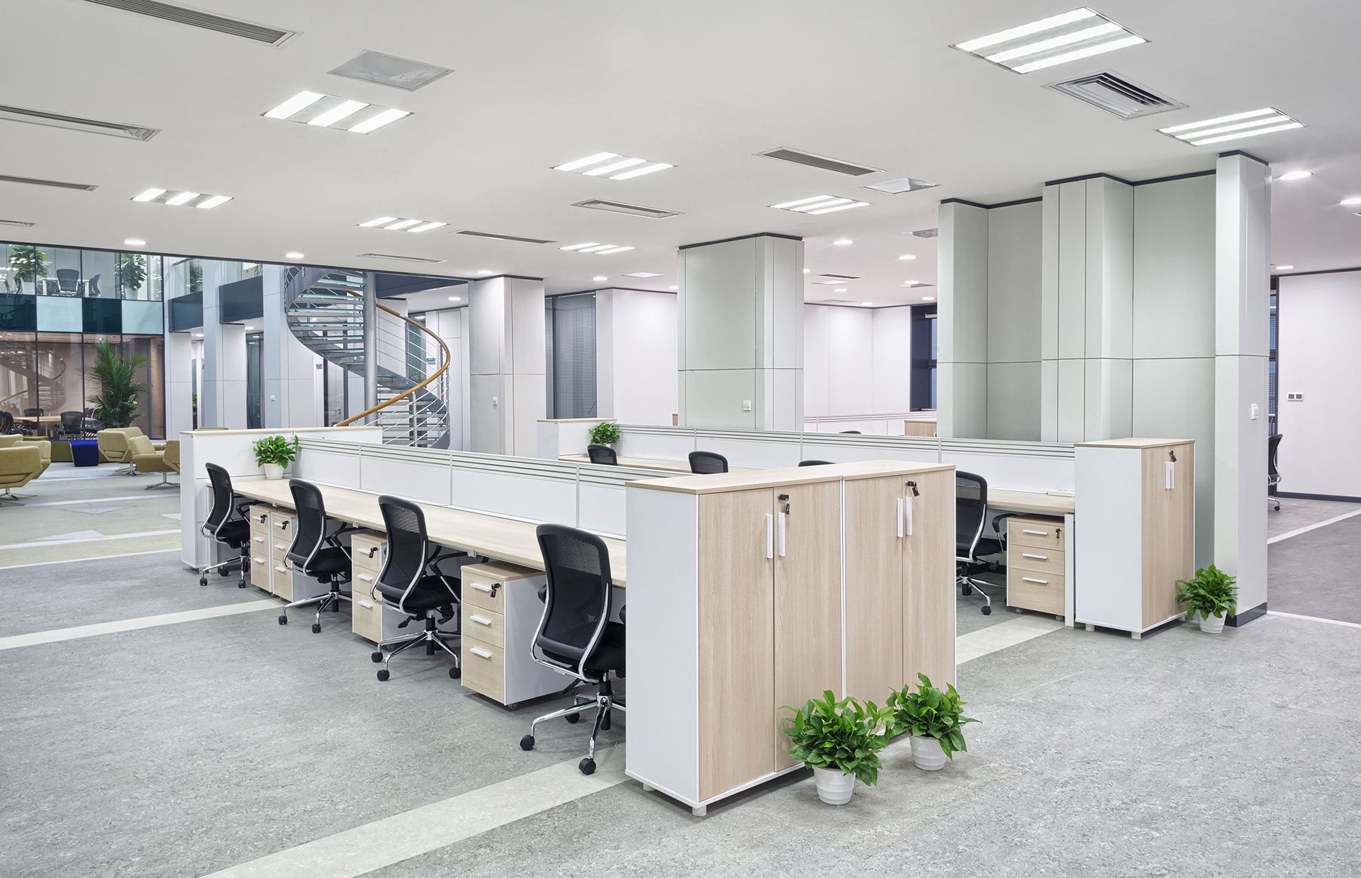 Bright modern open-plan office with desks, chairs, partitions, and potted plants under fluorescent lights
