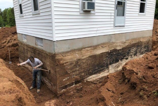 Person digging around a house foundation, showing exposed earth layers. White siding, air conditioner.