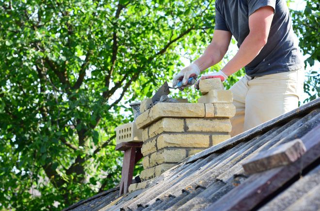 Person working on a brick chimney on a sloped roof with trees in the background