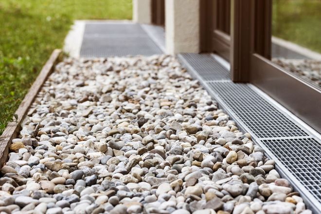 Gravel drainage bed beside a building with a metal grate and wooden posts