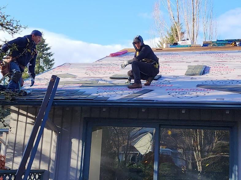 Two workers on a roof installing shingles, with one kneeling near the ridge and another standing by a ladder.