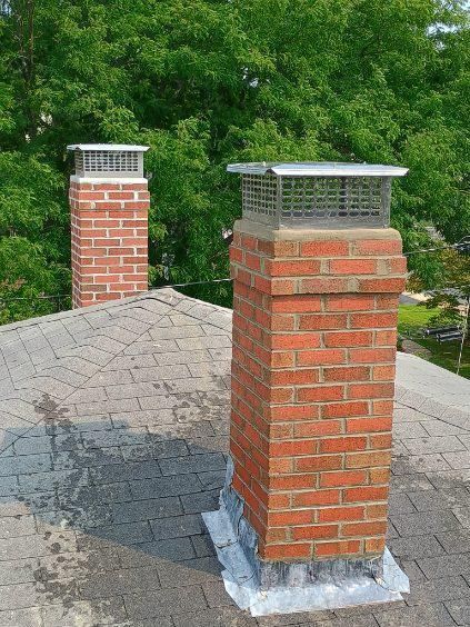 Two brick chimney stacks with metal caps on a shingled roof, with green trees in the background.