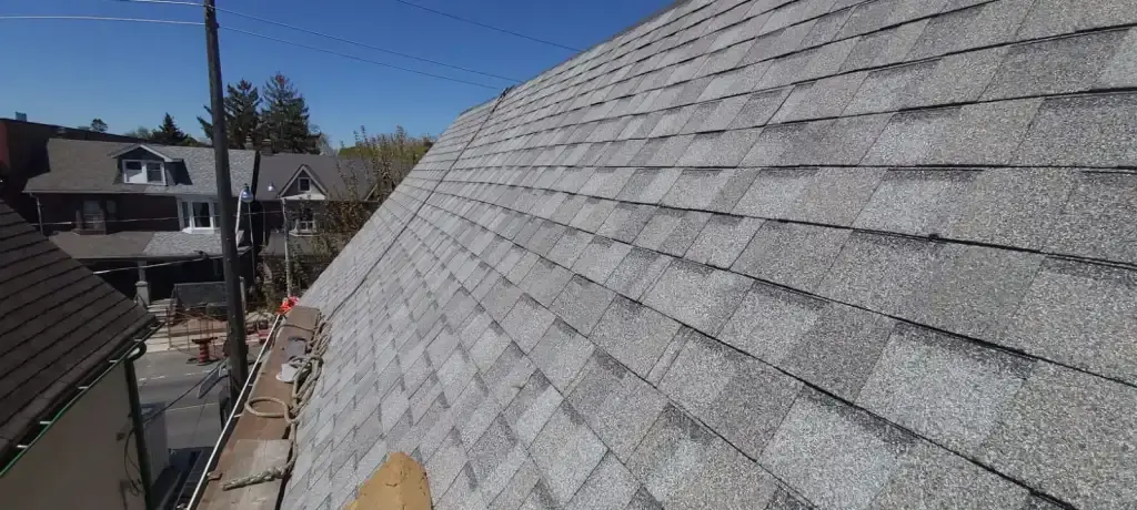 Steep gray shingled roof in sunlight with neighboring buildings and blue sky in the background