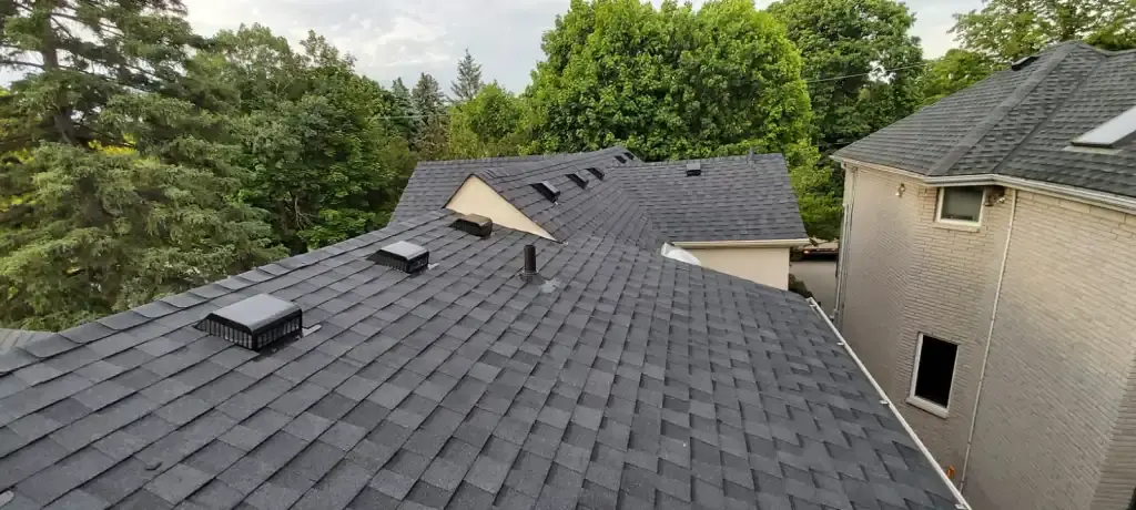 Roof with dark gray shingles, vents, and a damaged section beside a beige house and trees