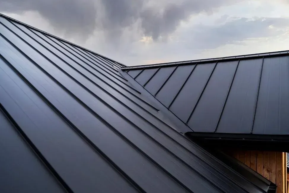 Dark gray metal roof with standing seams and a wooden wall under a cloudy sky