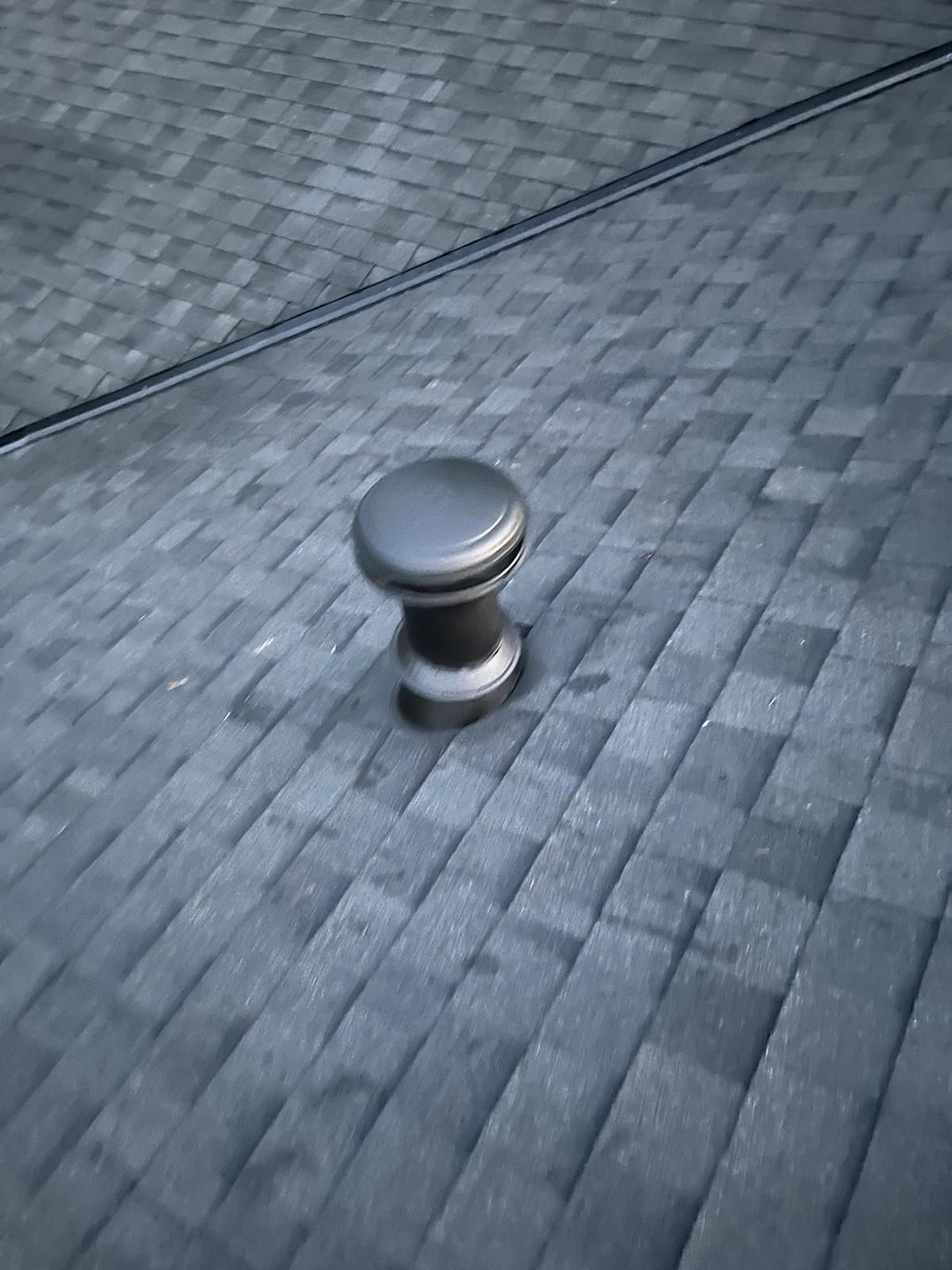 Metal vent cap on a gray shingled roof, viewed from above.