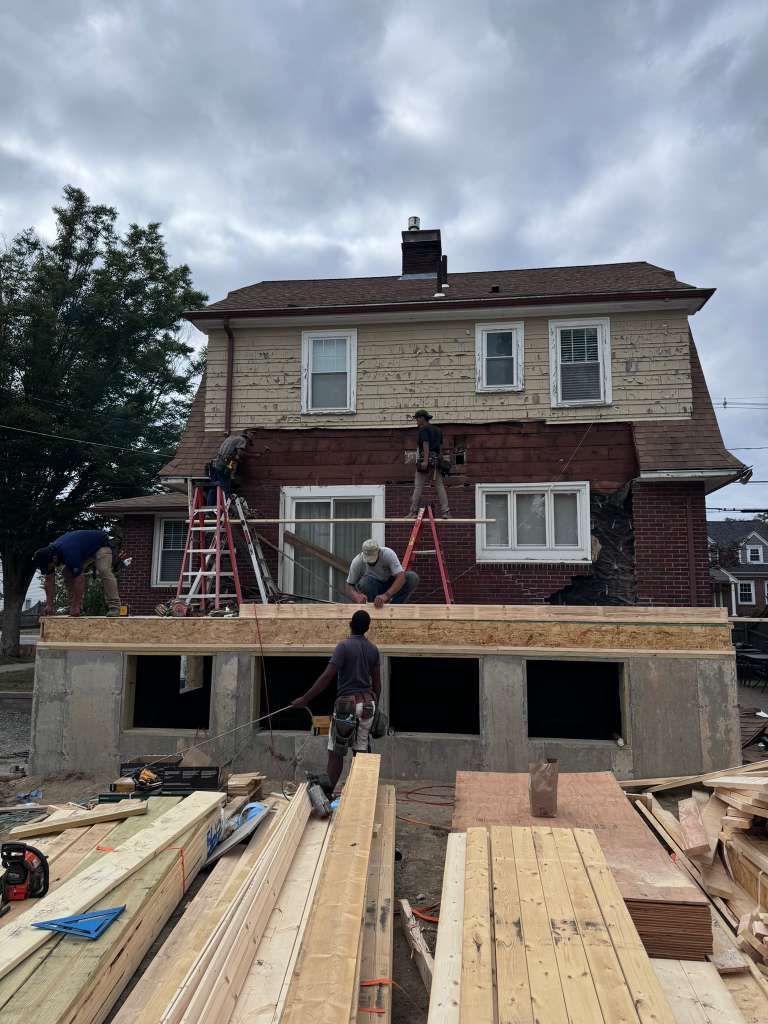 Workers framing an addition on a brick house, with lumber and ladders in front under a cloudy sky