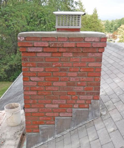 Red brick chimney with a metal cap on a gray shingled roof against trees.