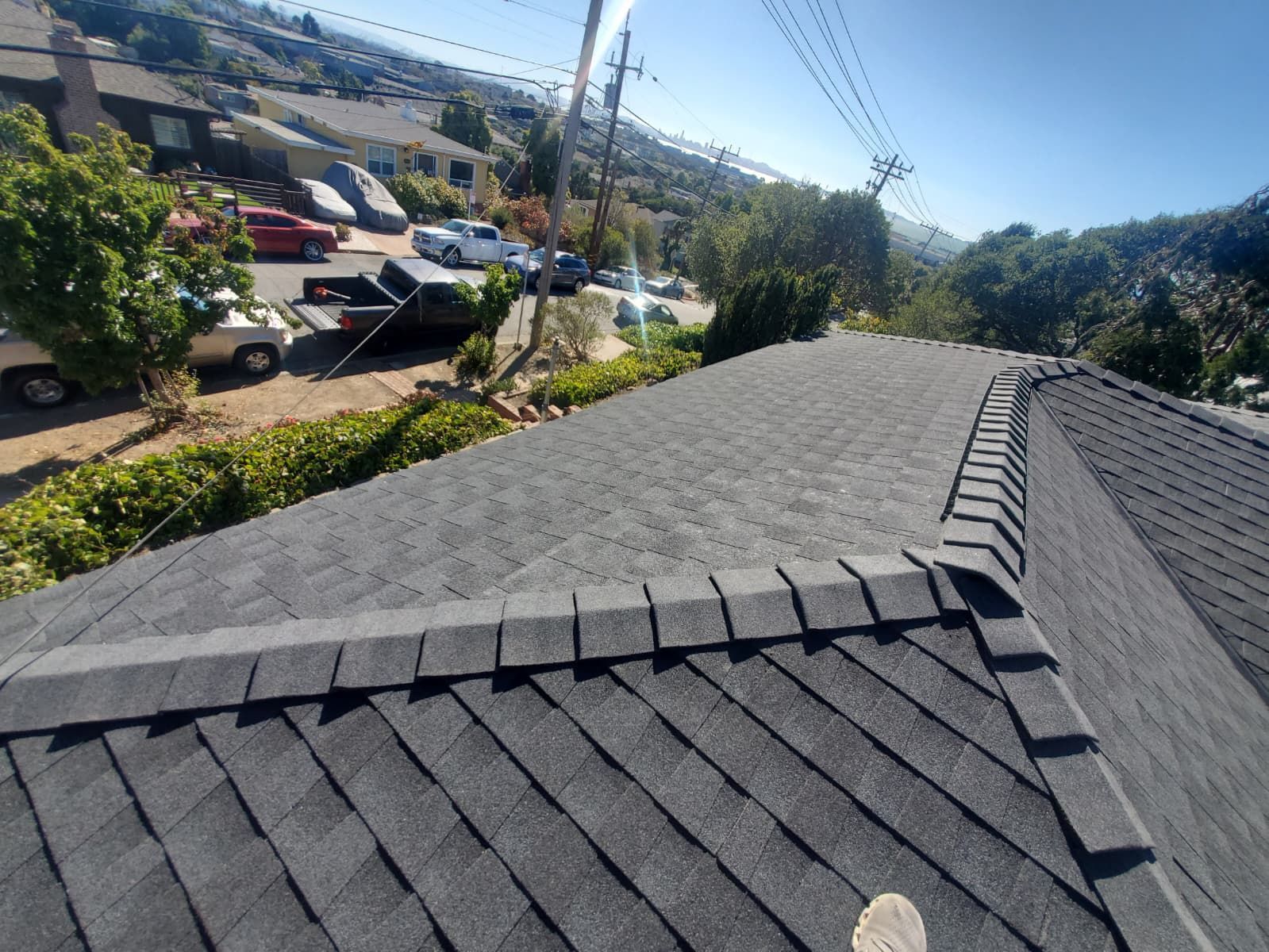 Rooftop view of dark gray shingles overlooking a sunny parking lot and hillside neighborhood