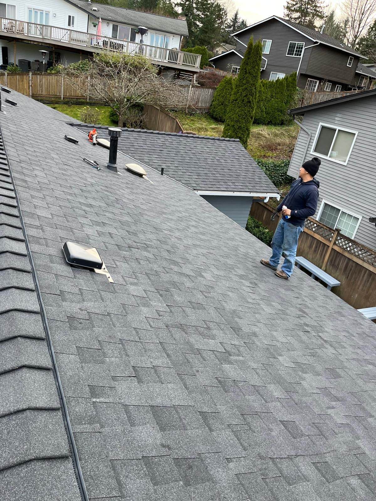 Person standing on a gray shingled roof beside a skylight in a residential neighborhood