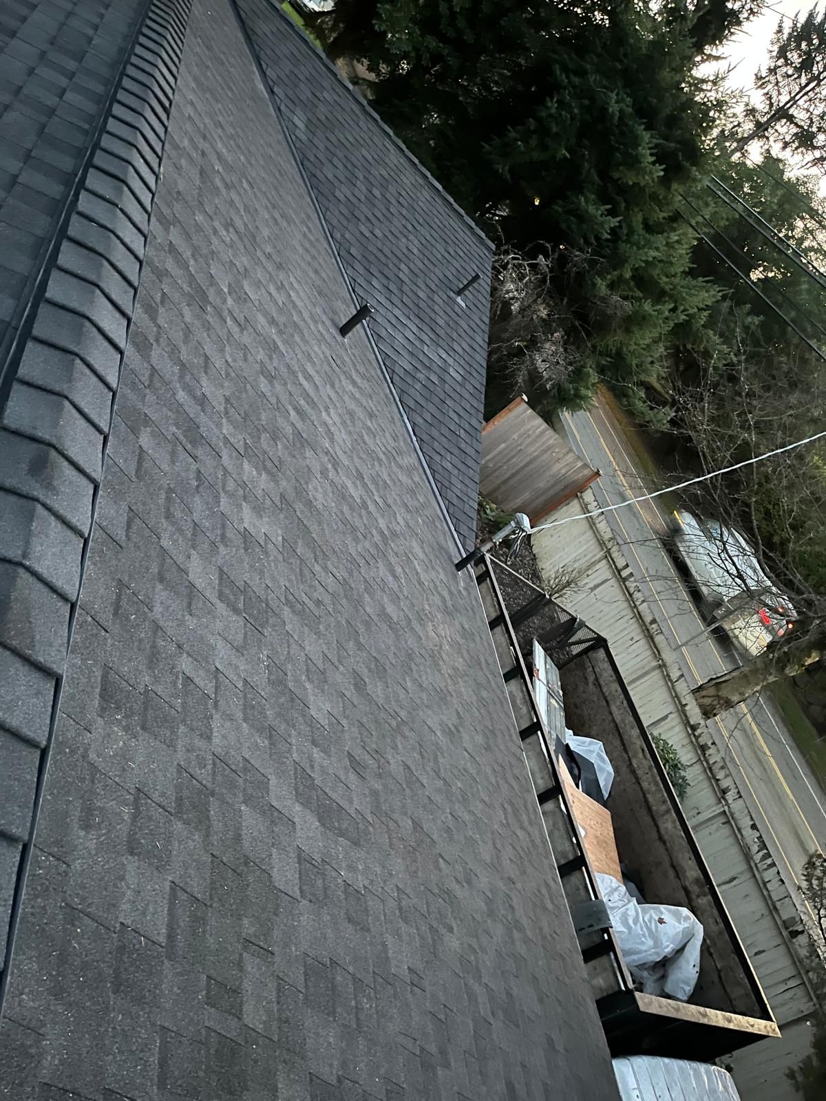 Tilted view of a gray shingled roof with a chimney and backyard deck below, lined by trees and a fence.