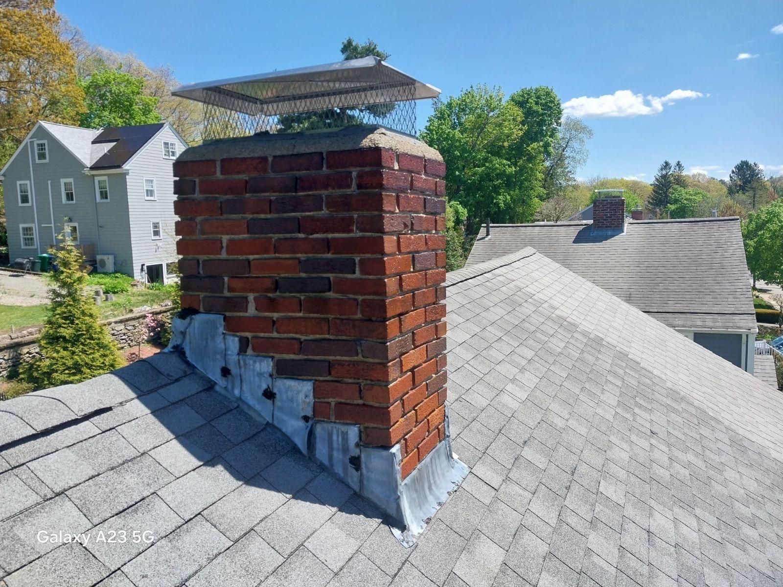 Brick chimney on a gray shingled roof with a metal cap, set against trees and nearby houses.