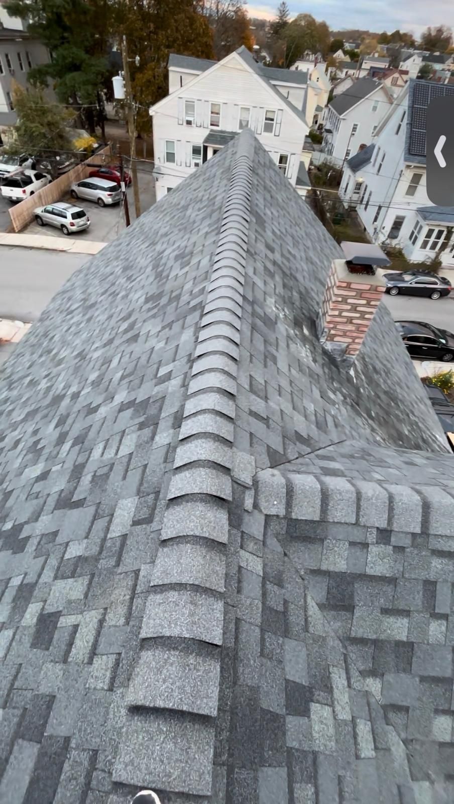 View from a gray shingle roof ridge overlooking houses, trees, and parked cars in a residential neighborhood
