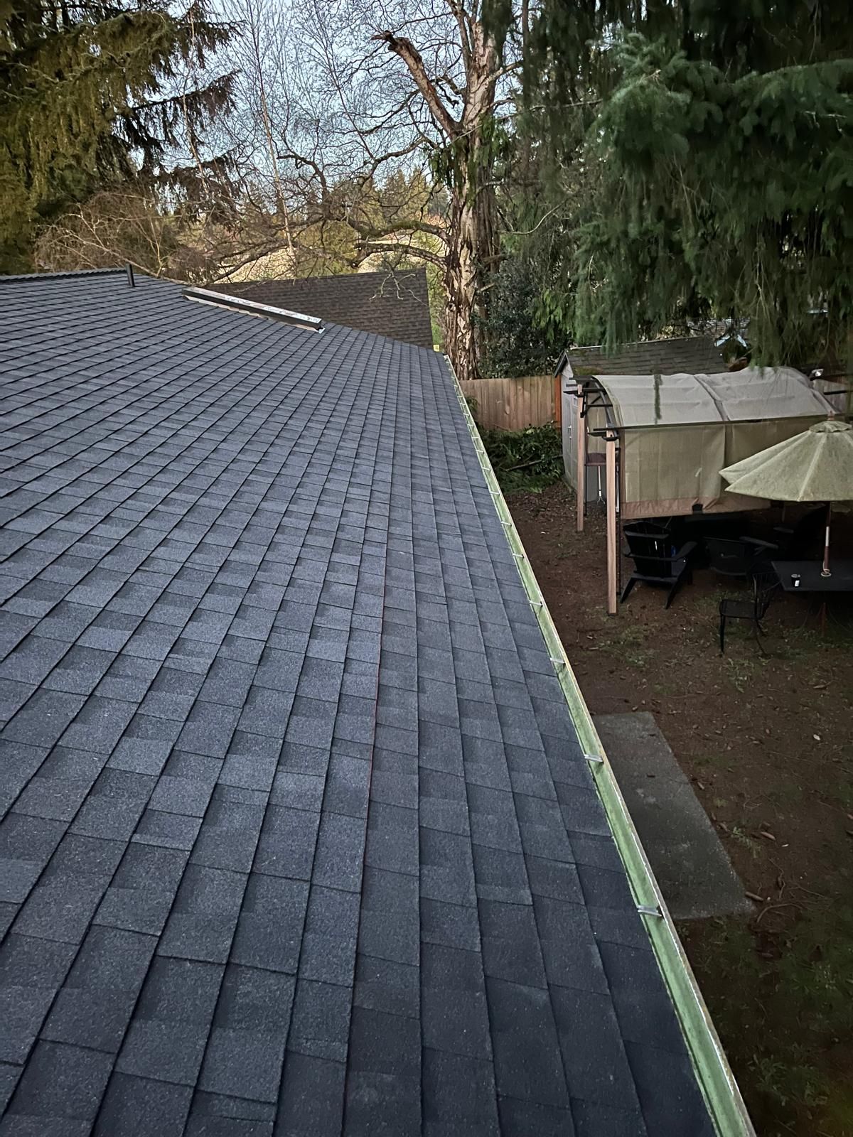 Shingled roof edge overlooking a backyard with trees, a patio table, and a covered canopy.