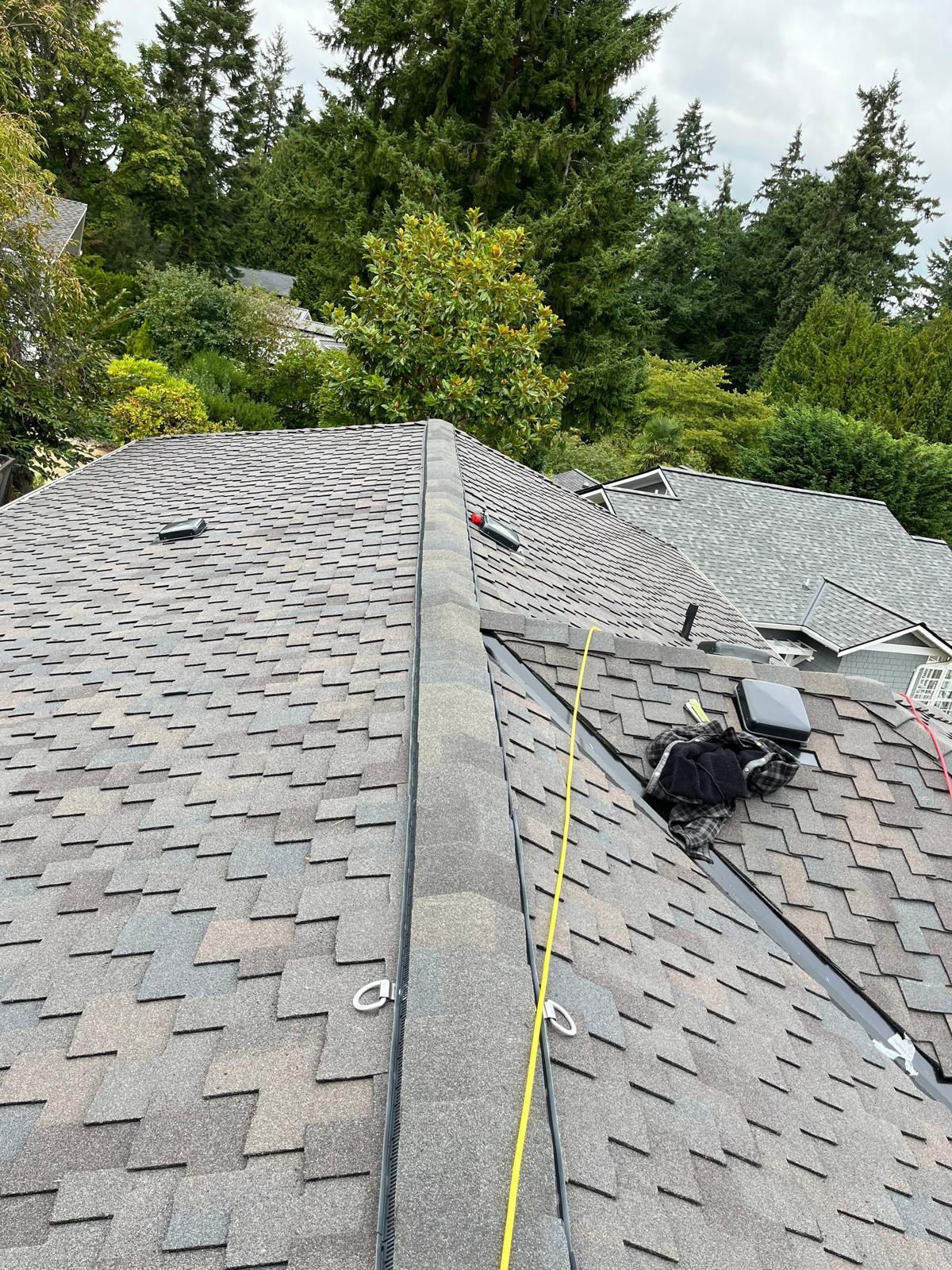 Roof under repair with gray shingles, tools, and a yellow safety line near a tree-lined house.