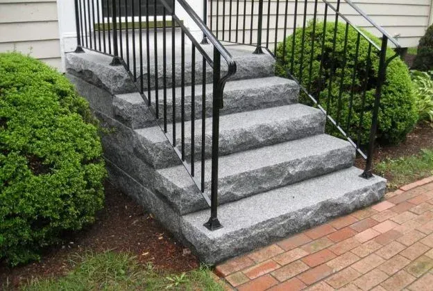 Granite front steps with black railing leading to a house entrance, bordered by shrubs and brick walkway