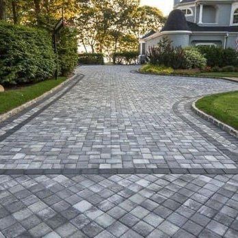 Stone driveway leading to a house, bordered by green lawns and trees at sunset.