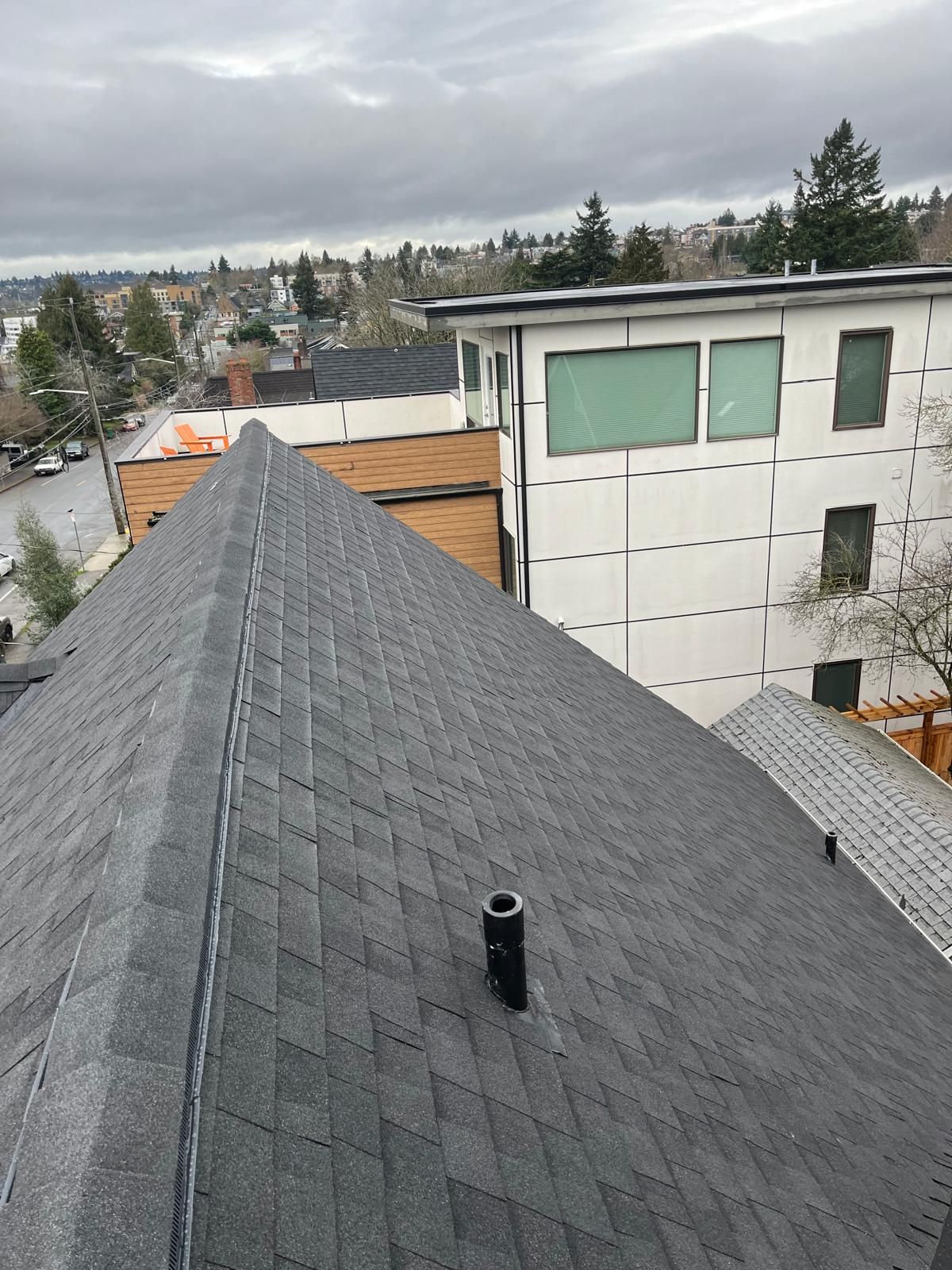 Rooftop with gray shingles and a vent, overlooking nearby houses under a cloudy sky.
