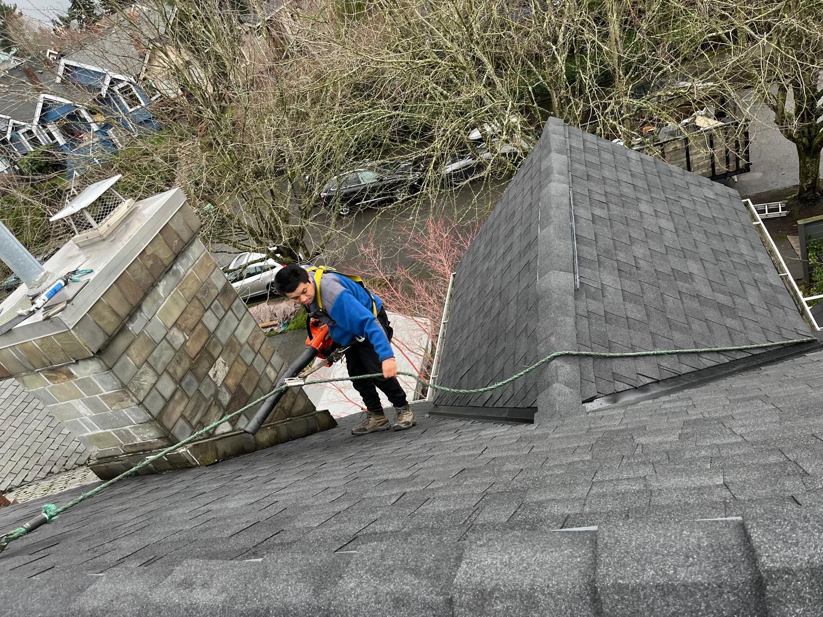 Worker on a steep shingled roof, securing a rope near a chimney in a wooded area.