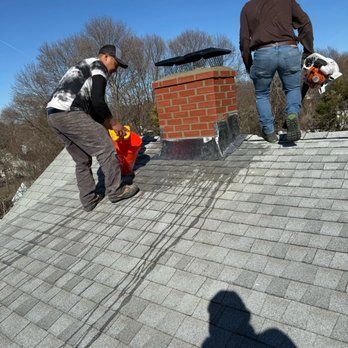 Workers repairing a shingled roof around a brick chimney on a sunny day